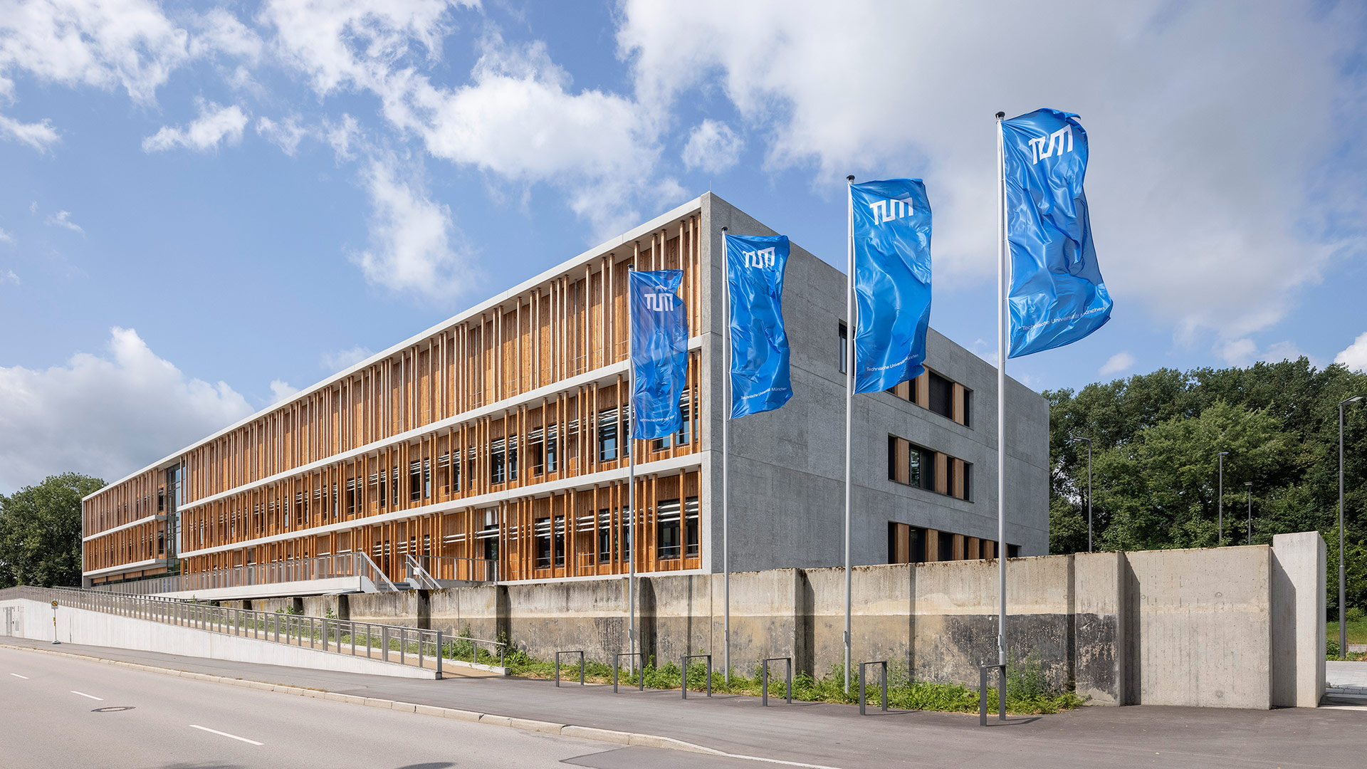 The building of Uferstraße 53, a large concrete cuboid with a wooden front. In front of it, four blue TUM flags are waving in the wind.