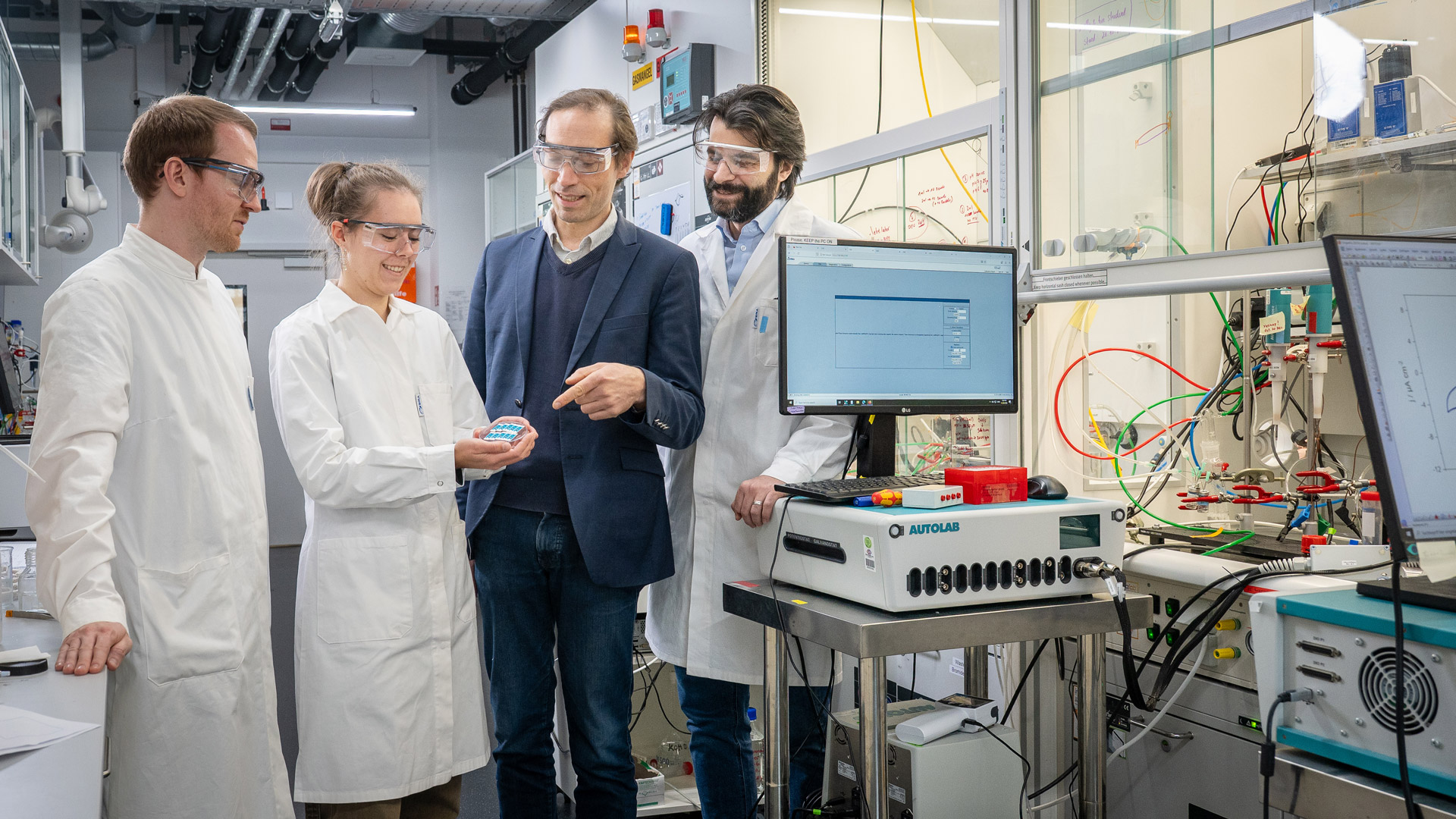 A woman and three men, one of them in suit, standing in a lab, looking at a batch of sensors. The sensors are small and of white and turqoise color.