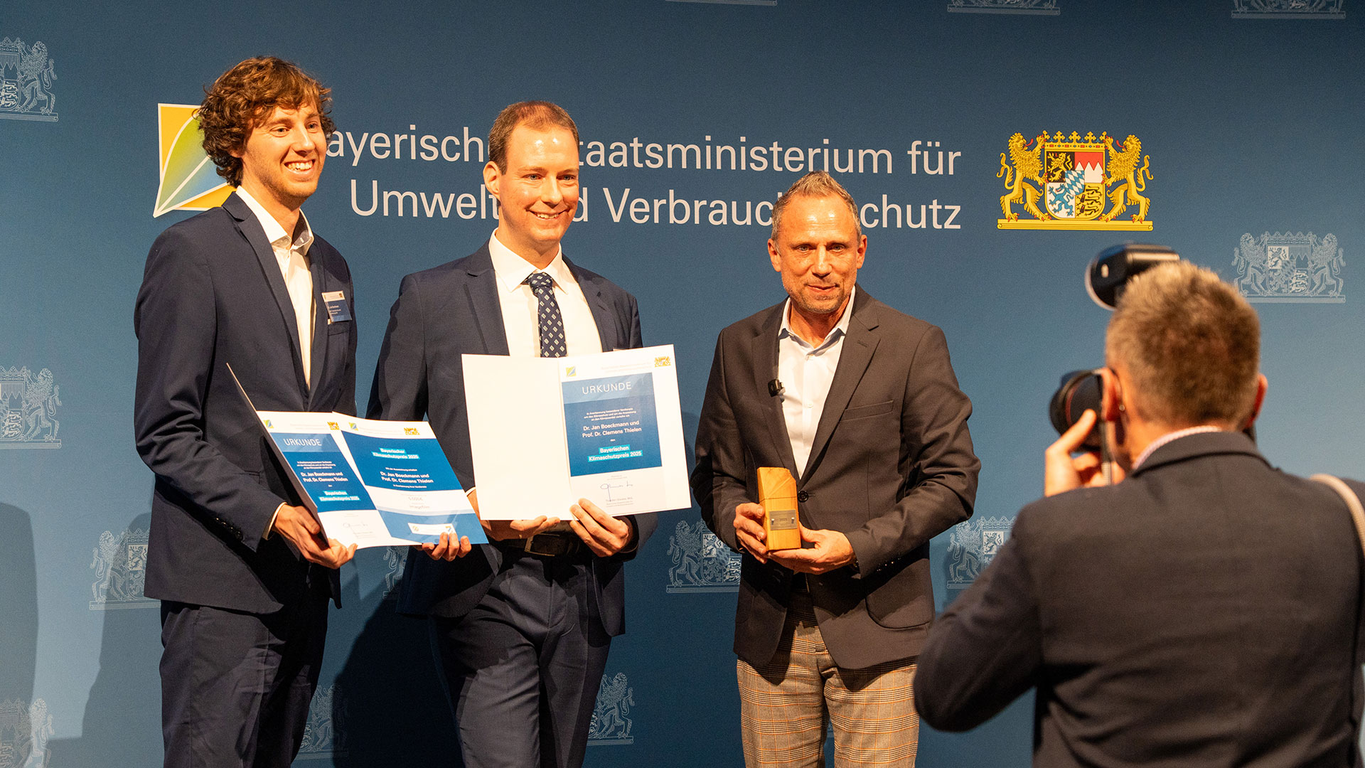 Three men in suits, standing side by side in front of a blue background decorated with bavarian state heralds, being photographed by a press photographer at the right of the picture. The two men on the left are presenting their award certificates whereas the third is holding the wooden trophy.
