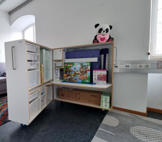 Eine geöffnete Sperrholzkiste auf Rädern, die ihren Inhalt offenbart: Spielzeug, ein Reisebett, Bücher, Buntstifte und vieles mehr.