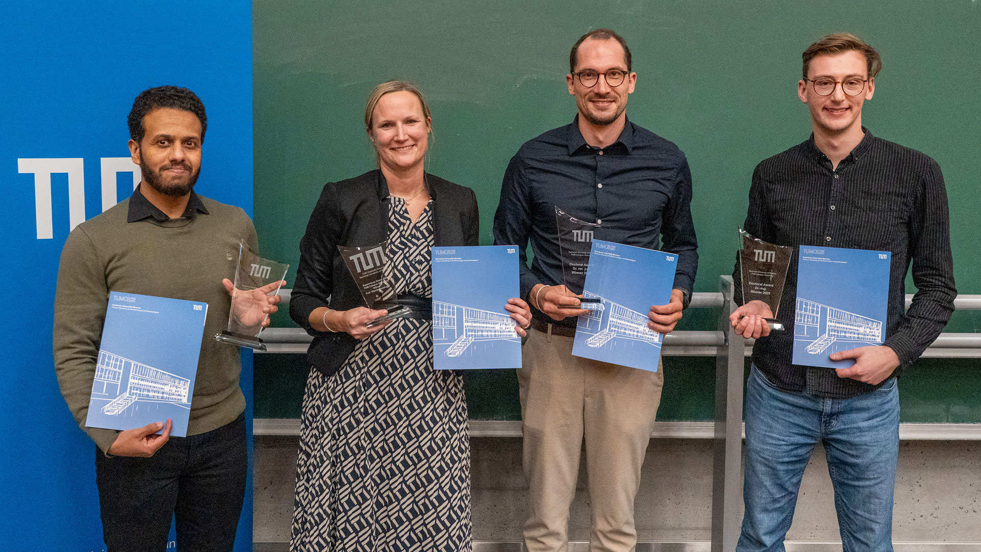 Four people, a woman and three men, in front of the board in lecture hall U7a, presenting their cerificates and trophys