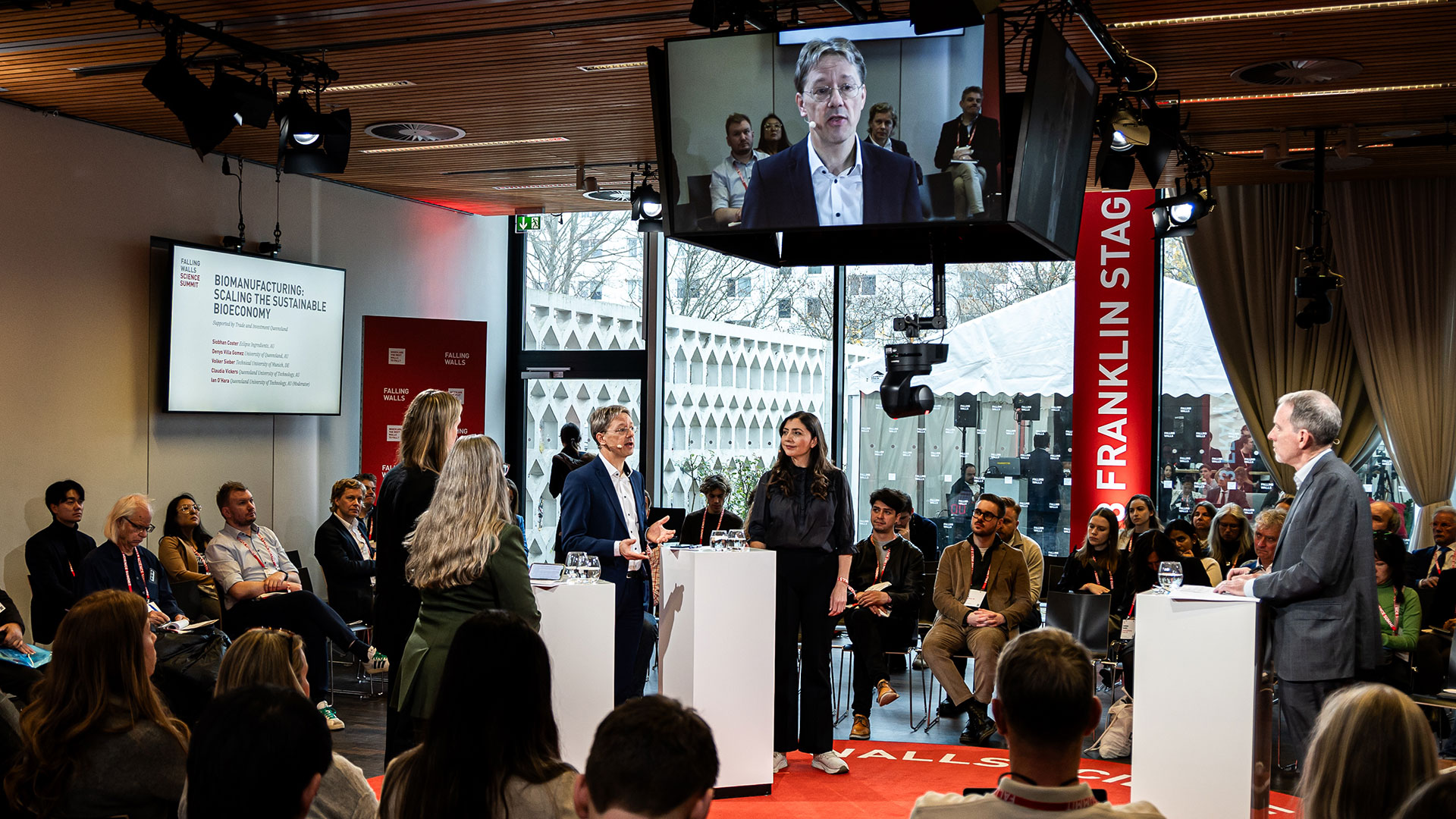 A circular, ground level stage in a room, with the audience sitting around it. On stage, a moderator on the right of the picture is facing three women and one man, Volker Sieber, on the left side of the frame. Above the stage is a cube with four live screens, showing a medium close-up of the speakering participant..