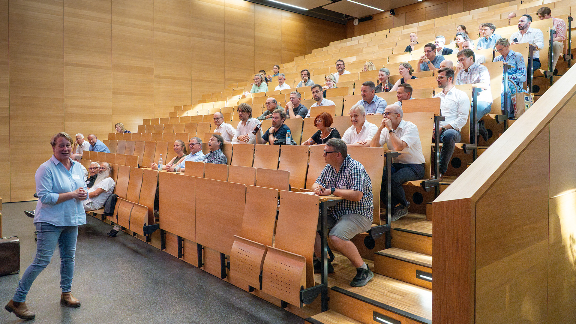 A group of people is sitting in the step auditorium. Prof. Anja Faße stand in front of them, delivering her welcome speech.