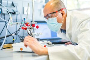 Ein Mann in Kittel und Maske untersucht einen Versuchsaufbau im Labor