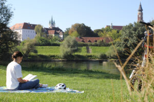 Eine Frau liest auf einer Decke im Grünen an der Donau ein Buch. Im Hintergrund die Stadt Straubing mit dem berühmten Stadtturm.