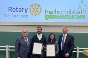 Vier Personen, zwei davon Preisträger des Wissenschaftspreises 2023 mit entsprechenden Zertifikaten, posieren vor der Tafel eines Hörsaals.