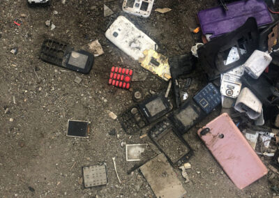 Teile kaputter Mobiltelefone am unbefestigten Boden, zum Teil bereits in den Sand eingetreten.